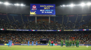 South Africa cricket team celebrates on the field after beating India by 76 runs at the Narendra Modi Stadium, with the giant stadium scoreboard displaying the final Super 8 match result.